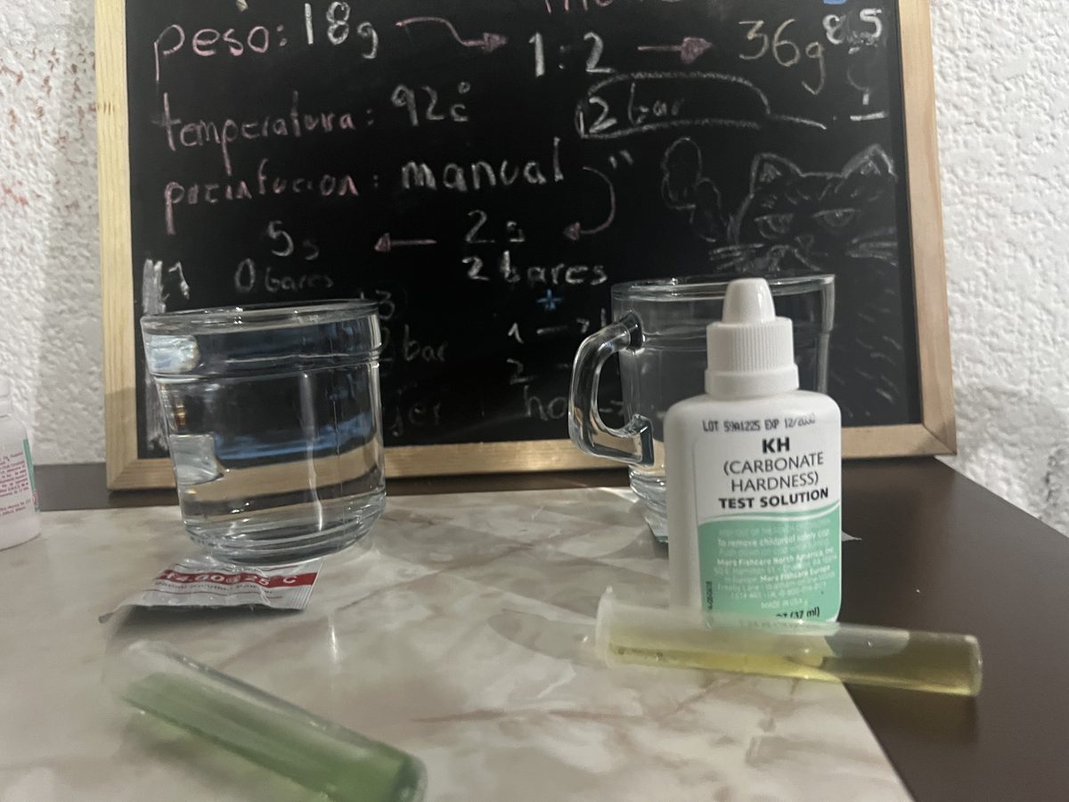 Complete water testing setup: glasses with water, test tubes with GH green and KH yellow results, KH reagent bottle, with notes chalkboard in background